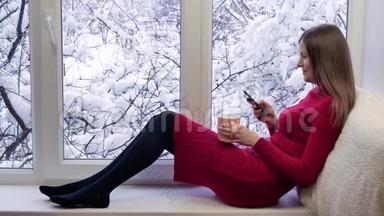 穿着红裙子的漂亮女孩坐在窗台上，喝茶，用智能手机。 冬天在外面。